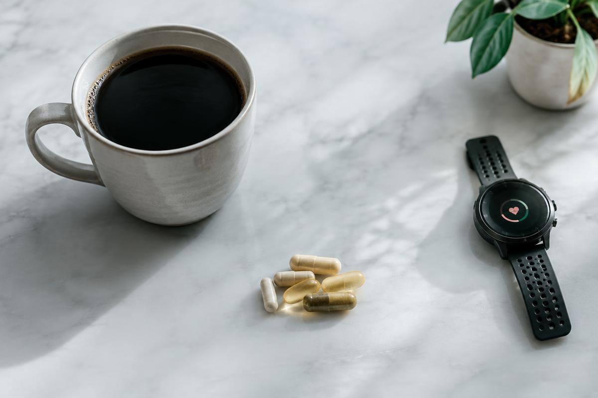 A cup of black coffee beside supplement capsules and a heart-rate watch on a marble counter