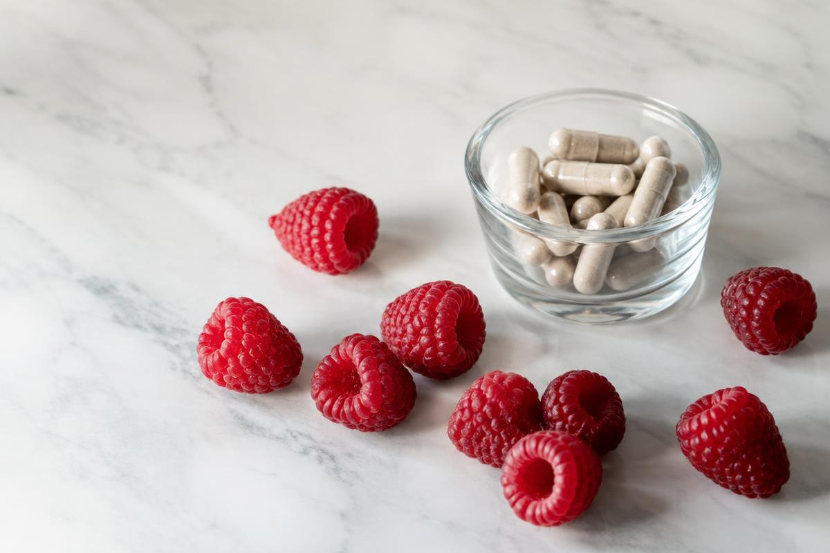 Fresh raspberries on a marble surface beside a glass dish of supplement capsules